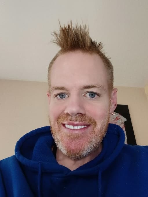 Man with a spiky reddish-blonde mohawk and beard, smiling in a blue hooded sweatshirt.